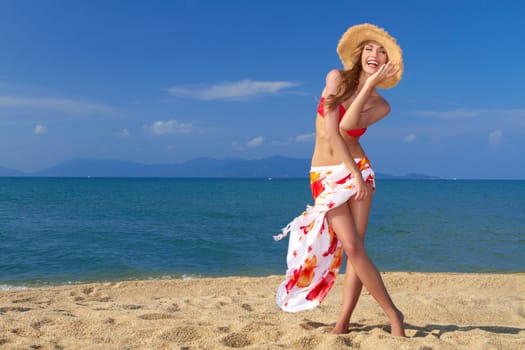Smiling woman in straw hat with scarf around her waist posing on sandy beach by the sea