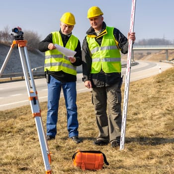 Two male geodesist with tacheometer checking plans standing by highway