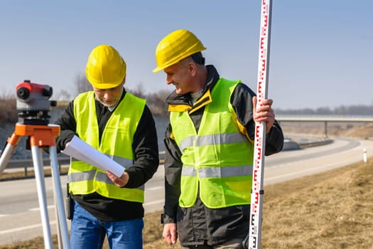 Two male geodesist with tacheometer checking plans standing by highway