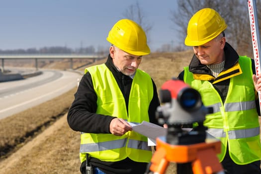Two male geodesist with tacheometer checking plans standing by highway