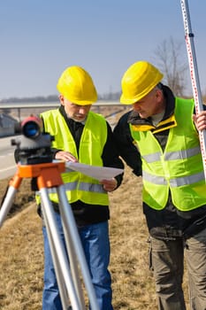 Two male geodesist with tacheometer checking plans standing by highway