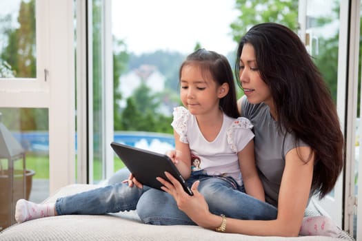 Cute little girl sitting with her mother on couch using a digital tablet.