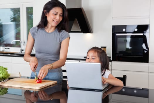 Cute girl using laptop while mother cuts vegetable in kitchen