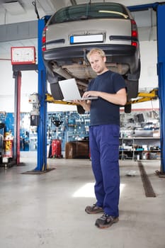 Full length portrait of smiling young mechanic using laptop in his auto repair shop