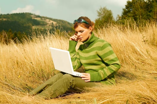 Young business woman working on laptop computer on golden meadow at mountains outdoors