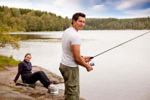 A man fishing on a lake with camping equipment and woman in background