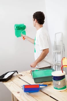 Casual man painting the wall of his new apartment with paint bucket in background