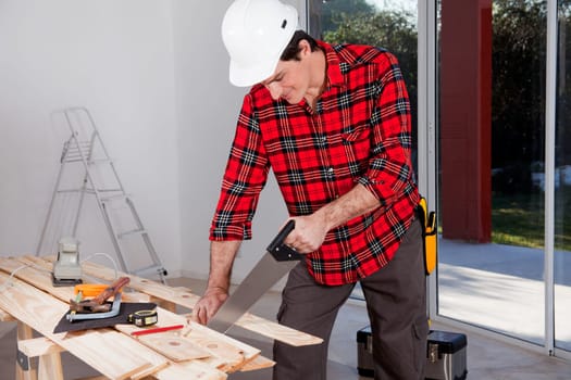 A construction worker using a hand wood saw while wearing a white safety helmet
