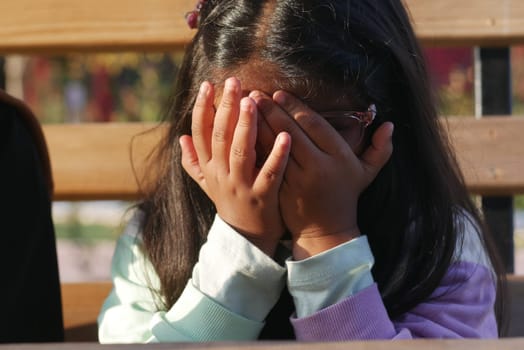 A young child hides her face with hands, expressing various emotions in a park.