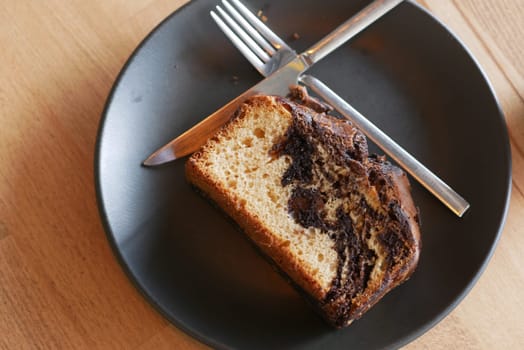 A slice of chocolate marble cake rests on a black plate with cutlery.