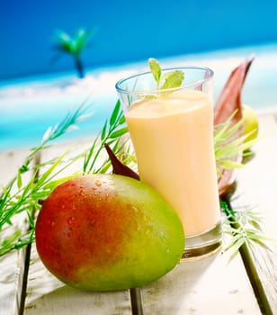 Glass of creamy mango and yoghurt smoothie dessert with a large ripe mango on a wooden deck overlooking the sea