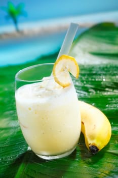Refreshing tropical banana smoothie and ripe yellow banana on a green frond with ocean backdrop