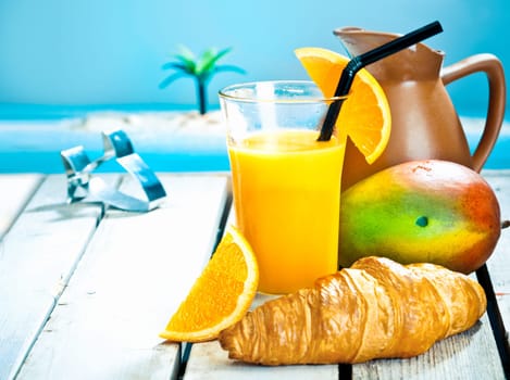 Tropical breakfast still life with a healthy mango and orange smoothie and a fresh ripe mango with a golden croissant against a blue ocean backdrop