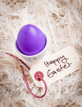 A vibrant purple Easter Egg in an egg cup among white feathers with a Happy Easter tag.