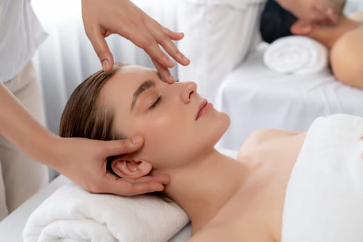 Caucasian couple enjoying relaxing anti-stress head massage and pampering facial beauty skin recreation leisure in dayspa modern light ambient at luxury resort or hotel spa salon. Quiescent