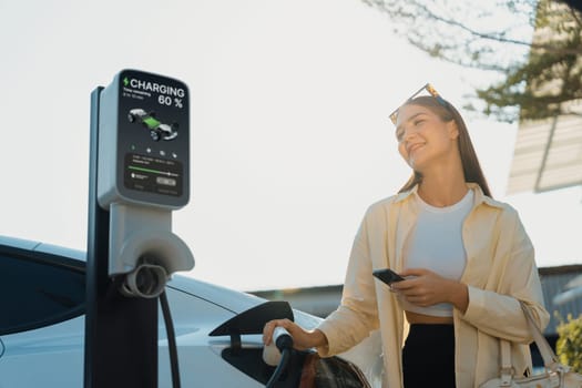 Young woman holding shopping bag and use smartphone to pay for electricity for recharging EV car battery from charging station at city mall parking lot. Modern woman go shopping by eco car. Expedient