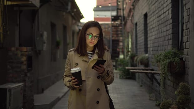 A young woman walks along a narrow street late at night and chats on her smartphone. A girl with glasses walks with a phone in her hands and drinks coffee. 4k