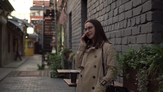 A young woman talks on the phone while standing against a wall on a narrow street. A girl with glasses and a coat speaks on the phone. 4k