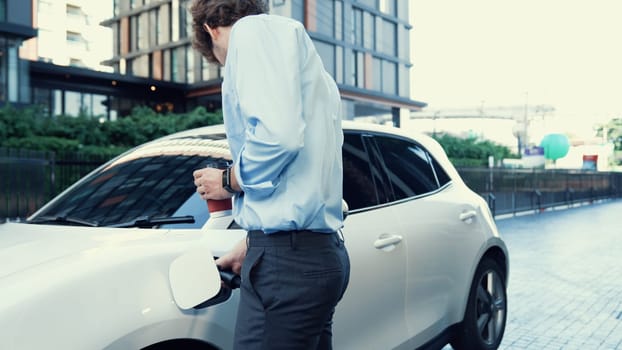 Suit-clad businessman with progressive ambition leaning on his electric vehicle while standing on a charging station with a power cable plug and a renewable energy-powered electric vehicle.