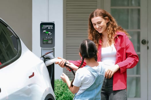 Happy little young girl learn about eco-friendly and energy sustainability as she help her mother recharge electric vehicle from home EV charging station. EV car and modern family concept. Synchronos