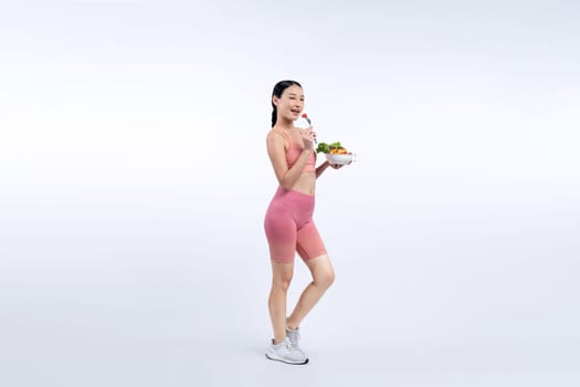 Young sporty Asian woman in sportswear holding salad bowl fill with vibrant of fruit and vegetable. Natural youthful and fit body lifestyle with balance nutrition on isolated background. Vigorous