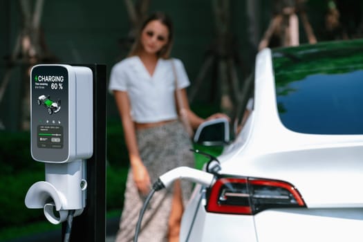 Young woman travel with EV electric car charging in green sustainable city outdoor garden in summer. Urban sustainability lifestyle by green clean rechargeable energy of electric BEV vehicle innards