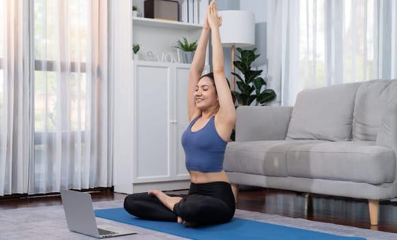 Asian woman in sportswear doing yoga exercise on fitness mat as her home workout training routine. Healthy body care lifestyle woman watching online yoga video on laptop. Vigorous