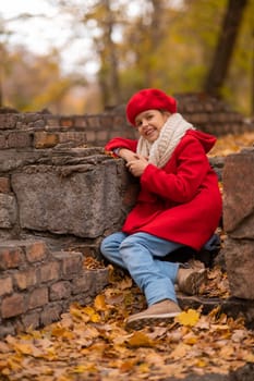 Smiling caucasian girl in a red coat and beret sits on a brick wall on a walk in autumn