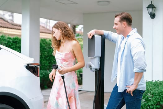 Happy and lovely couple with eco-friendly conscious recharging electric vehicle from EV home charging station. EV car technology utilized for residential home to future sustainability. Synchronos