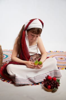 Portrait of Little girl in a stylized Tatar national costume with berries and a brush of grapes on a white background in the studio. Photo shoot of funny young teenager who is not a professional model