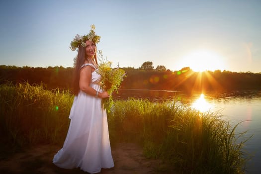 Adult mature brunette woman in a white dress, sundress and a wreath of flowers in summer by the water of river or lake in evening at sunset. Celebration of the Slavic pagan holiday of Ivan Kupala