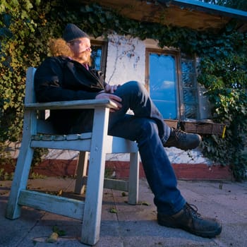 Full length view of an elderly man in a knitted cap sitting on a chair in front of his gazebo and looking into the distance in oblique,sideways and deep view at dusk