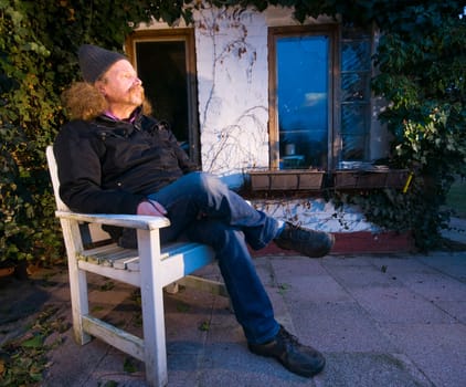Full length view of an elderly man in a knitted cap sitting on a chair in front of his gazebo and looking into the distance in oblique,sideways and deep view at dusk