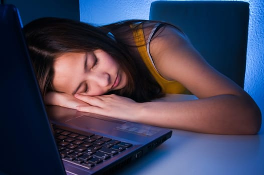 Head and shoulders shot of a young black-haired woman with her head resting on her hands in front of the computer keyboard sleeping in wide-angle and sideways view at table height