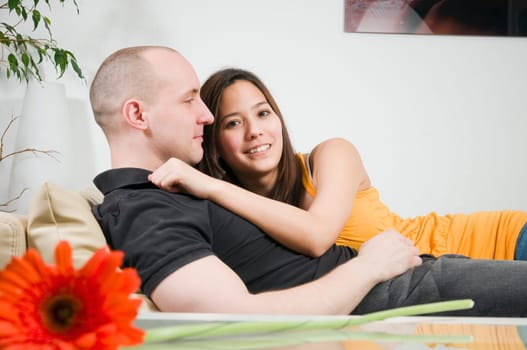 Upper body portrait of a young couple at home lying arm in arm on the couch with a single flower lying in front of it on the table