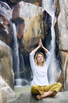Young woman doing yoga exercises over waterfall background.