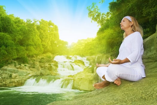 Young woman meditating at sunrise time near waterfall