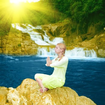 Young woman meditating at sunrise time near waterfall