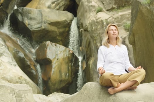 Young woman doing yoga exercises over waterfall background.