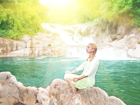 Young woman meditating at sunrise time near waterfall