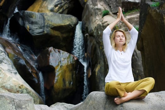 Young woman doing yoga exercises over waterfall background.