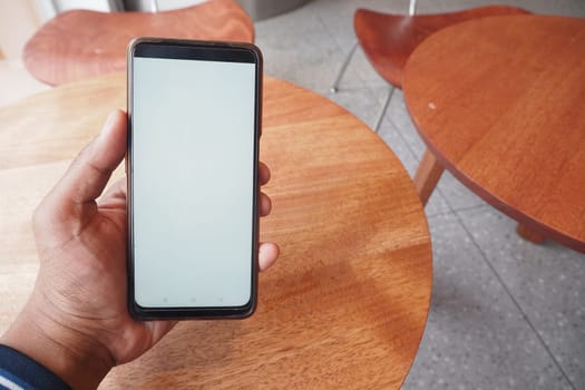 Individual holds a smartphone with a blank screen at a wooden table.
