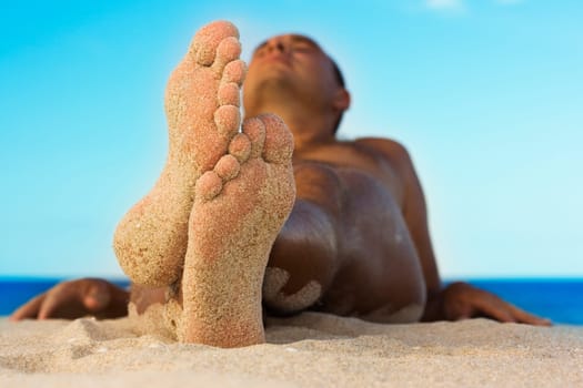Man relaxing on the tropical beach. Focus on his feet