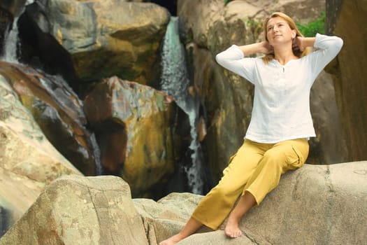 Young woman relaxing on the stone over waterfall background