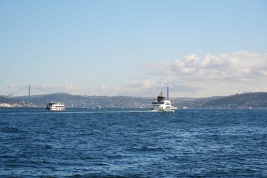 Two boats travel across the blue waters near Istanbul with a bridge in view.