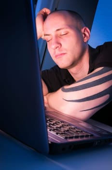 Head and shoulders shot of young man sleeping in front of laptop with head resting on hands against blue night background