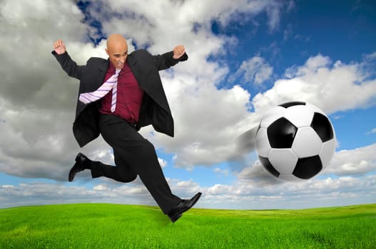 Businessman in a acrobatic pose kicking a soccer ball in a green field
