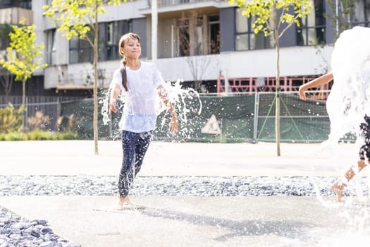 Cheerful young teen girl in city fountain, girl in wet clothes is having fun and enjoying the cool summer water, background city architecture. High quality photo