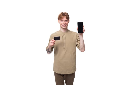 young well-groomed caucasian man with red hair dressed in a khaki shirt and brown trousers holds a credit card and a smartphone with a mockup.