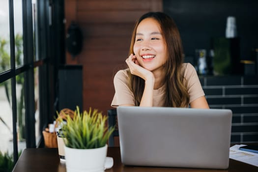 Happy Asian young businesswoman sitting alone at cafe desk with laptop computer she looking out of window, charming woman working outside and smiling in coffee shop freelancer thinking about new ideas